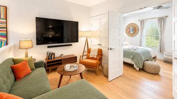 A modern living room with a flat-screen TV, a green sofa, a wooden coffee table, and a bed with a green blanket.