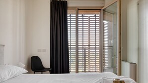Hotellihuone, jossa on sänky, tuoli, ikkuna, jossa on sälekaihtimet, ja ovi.