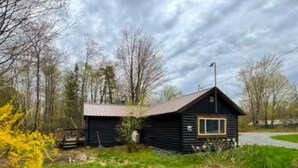 Eine kleine Holzhütte mit geneigtem Dach, umgeben von Bäumen und einer Wiese.