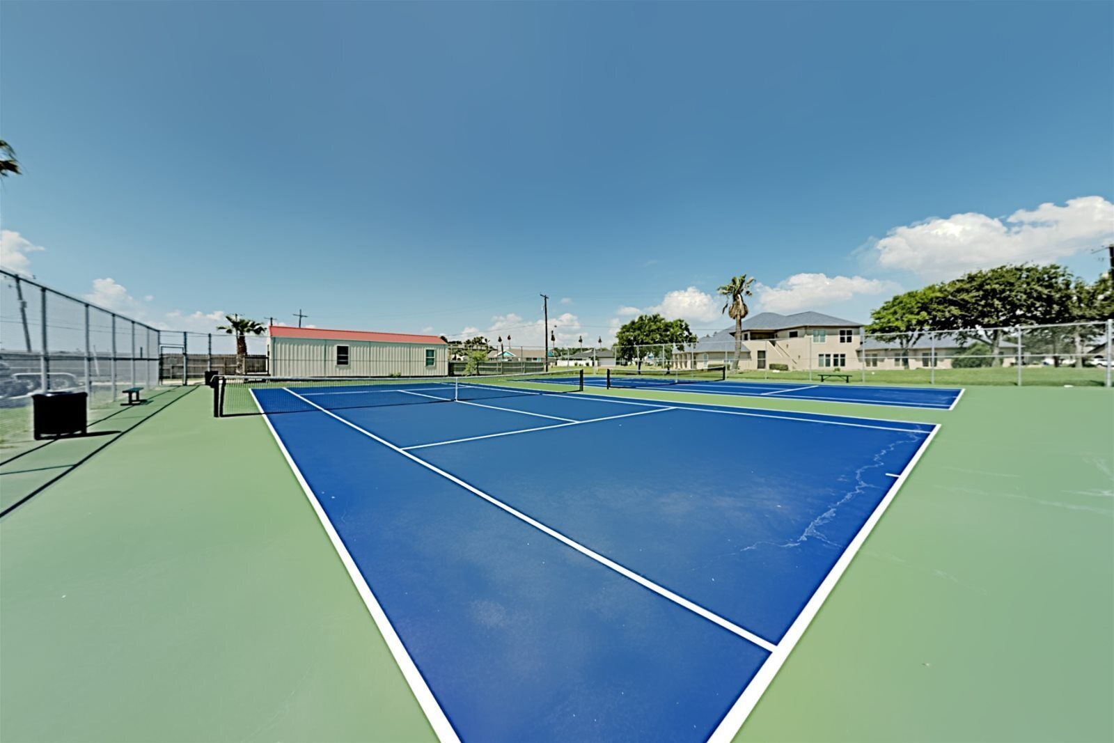 Una cancha de tenis exterior con red, rodeada por una valla y edificios al fondo.
