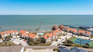 Een kustgebied met woonhuizen, meerdere tweelaagse woningen, een steiger die in zee reikt, en een tennisbaan.