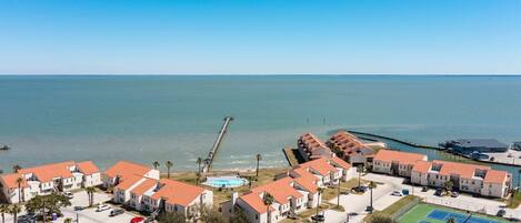 Un área residencial costera con varias casas de dos pisos, un muelle que se adentra en el mar y una cancha de tenis.