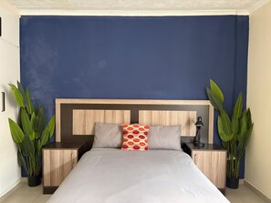 Een bed met een houten hoofdbord, twee nachtkastjes, een lamp en twee potten met planten.