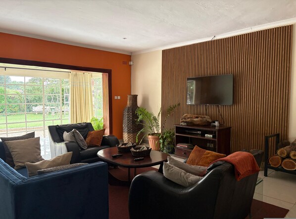 Een woonkamer met een bank, fauteuils, een salontafel, een televisie en een groot raam met gordijnen.