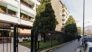 Een omheind woongebied met een verharde looppad, een hoge naaldboom en een meerlaags gebouw met balkons.