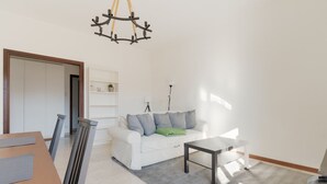 Een woonkamer met een witte bank, een bijzettafeltje, een kroonluchter en een boekenplank.