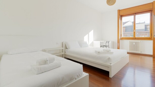 Een slaapkamer met twee bedden, een houten vloer, een groot raam en een hanglamp.