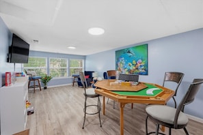 A room with a wooden table, chairs, a painting, and a television on the wall.