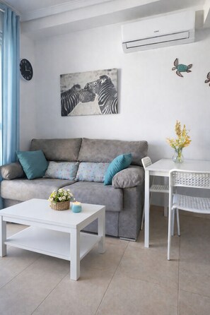 Ein Wohnzimmer mit grauem Sofa, weißem Couchtisch und einem kleinen Tisch mit einem Stuhl. An der Wand ist eine Klimaanlage angebracht, und es hängt ein Zebra-Gemälde.