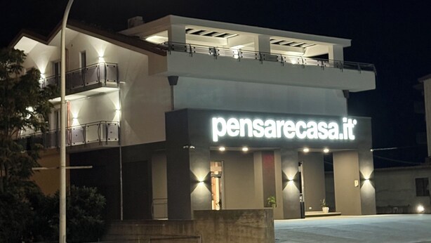 Un bâtiment moderne, sur lequel on peut voir, la nuit, une enseigne bien visible avec l’inscription « pensarecasa.it ».