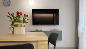 Un salon moderne avec une table à manger en bois, une chaise noire, un téléviseur fixé au mur et un vase de fleurs.