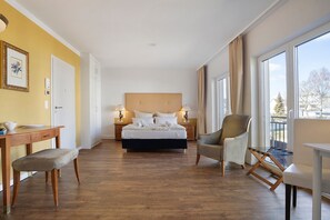 Ett hotellrum med en säng, ett skrivbord, en stol, ett litet bord och stora fönster.