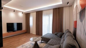 Un soggiorno moderno con una TV a schermo piatto, un divano componibile, un tavolino da caffè e un'ampia finestra con tende.