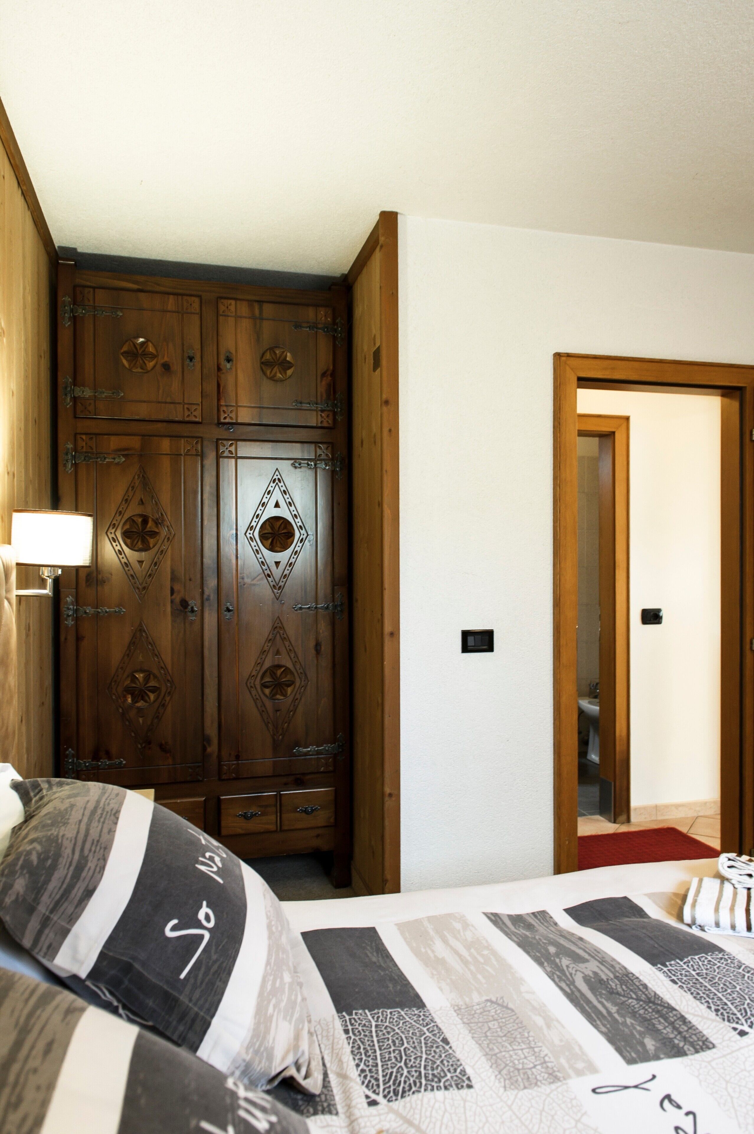 A bedroom with a wooden wardrobe, a bed with patterned bedding, and a nightstand with a lamp.