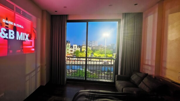 A hotel room with a large window, a sofa, and a wall-mounted screen displaying 'DARK & CHILL R&B MIX'. - Klara House Phuc Dong (Hanoi)