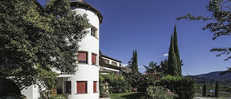 Un immeuble à plusieurs étages, doté d’une tour circulaire, entouré d’une végétation luxuriante et surplombé d’un ciel bleu azur.