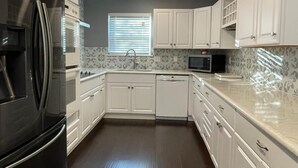 Una cocina con armarios blancos, un refrigerador negro, un microondas y una ventana con persianas.