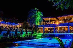 Zona de piscina con palmera, mobiliario exterior y un edificio con restaurante.
