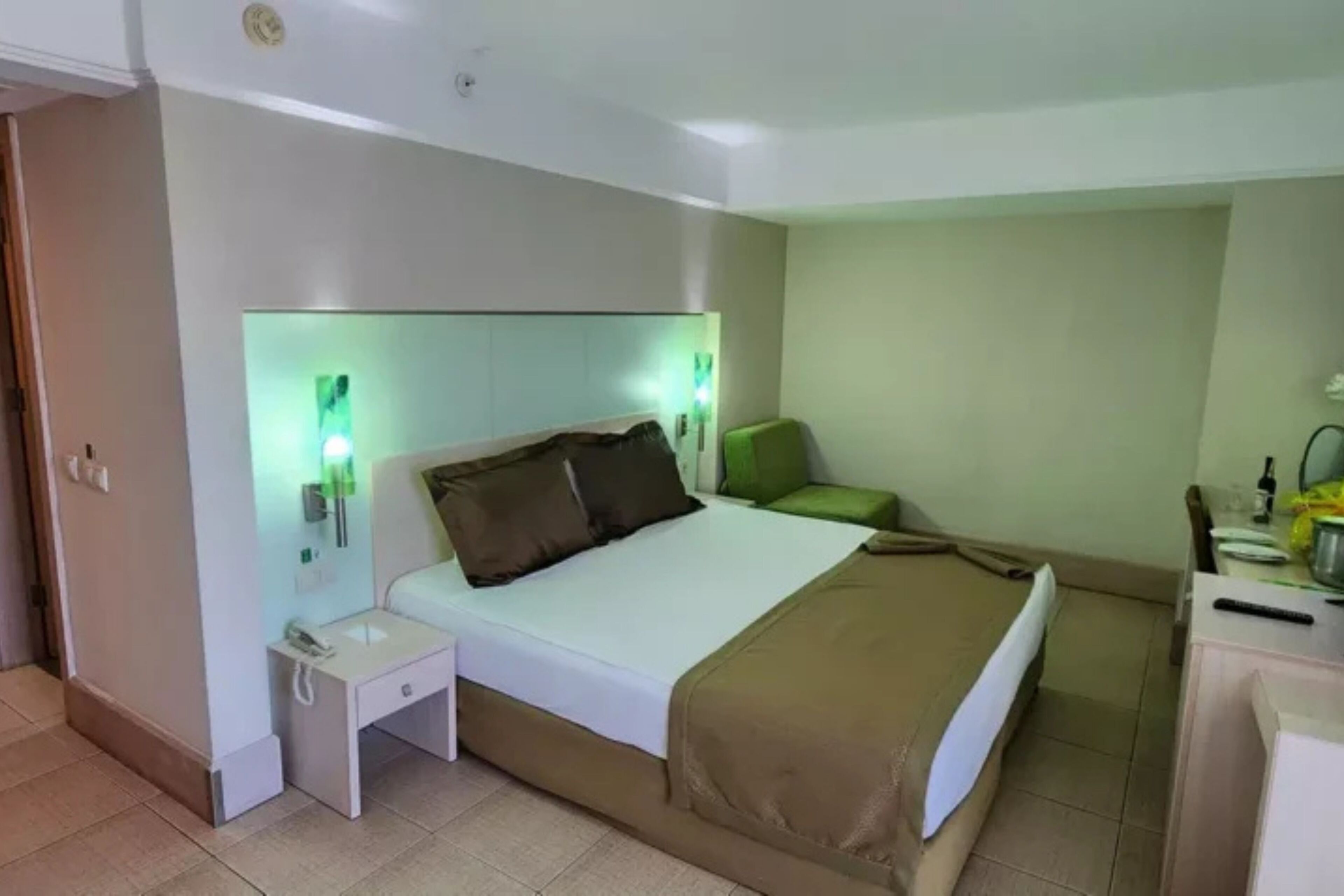 Une chambre d’hôtel avec un lit, une table de chevet, un fauteuil vert et un bureau avec une lampe.