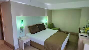 Une chambre d’hôtel avec un lit, une table de chevet, un fauteuil vert et un bureau avec une lampe.