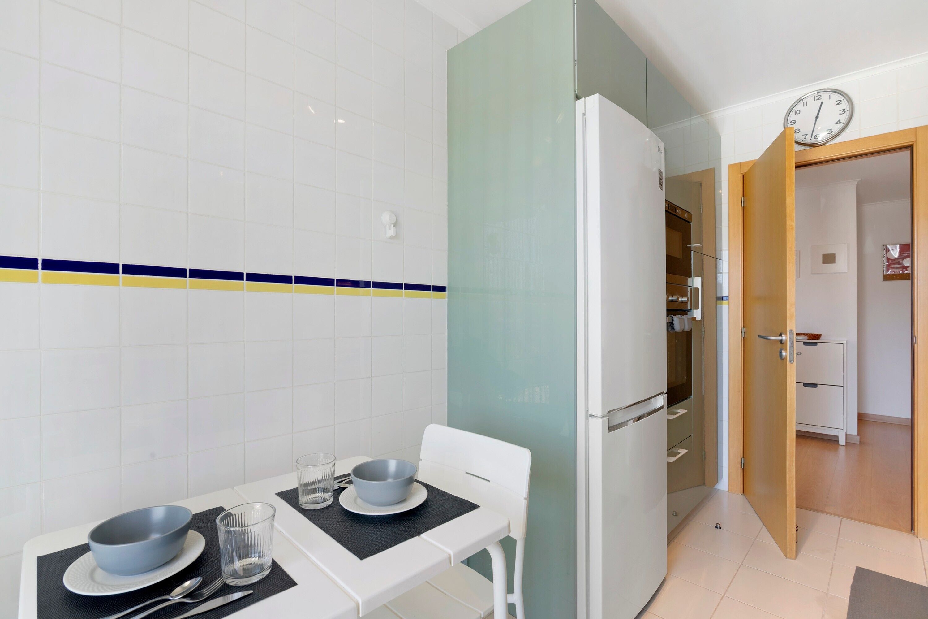 Una cocina pequeña con un refrigerador, una mesa con un tazón y un vaso, y un reloj en la pared.