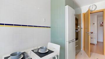 Una cocina pequeña con un refrigerador, una mesa con un tazón y un vaso, y un reloj en la pared.