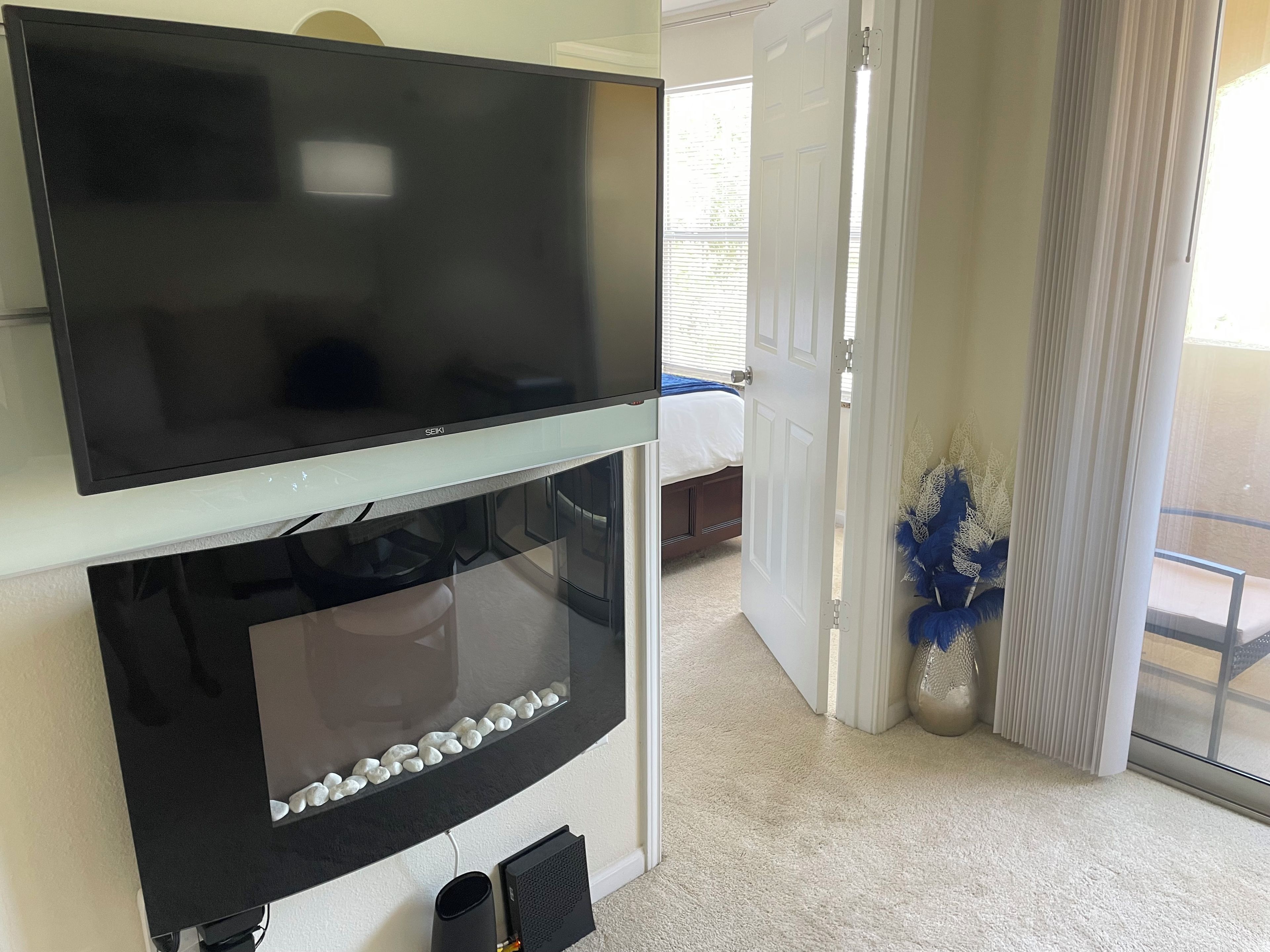 A flat-screen TV mounted above a fireplace with a stone border, a door leading to a balcony, and a potted plant with blue foliage.
