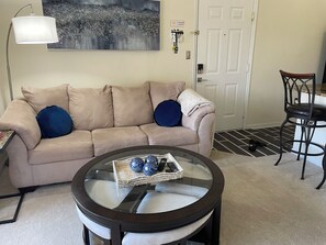 Ein Wohnzimmer mit einem beigefarbenen Sofa, zwei blauen Kissen, einem Glastisch mit Dekoration und einer weißen Lampe, sowie einer Tür mit Schlüsselkartenleser.