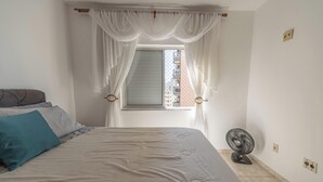 Ein Schlafzimmer mit einem Bett, einem Fenster mit Vorhängen, einem Ventilator und Blick auf Gebäude.