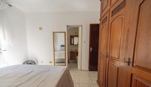 A bedroom with a bed, a wooden wardrobe, a mirror, and a door leading to a bathroom. - Apto em Praia Grande Frente a Praia da Guilhermina (Praia Grande)