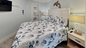 A bedroom with a bed, a nightstand, a lamp, a wall-mounted TV, a door, and a wall decoration. - Victorian 9124 - Good Times & Tan Lines (Galveston)
