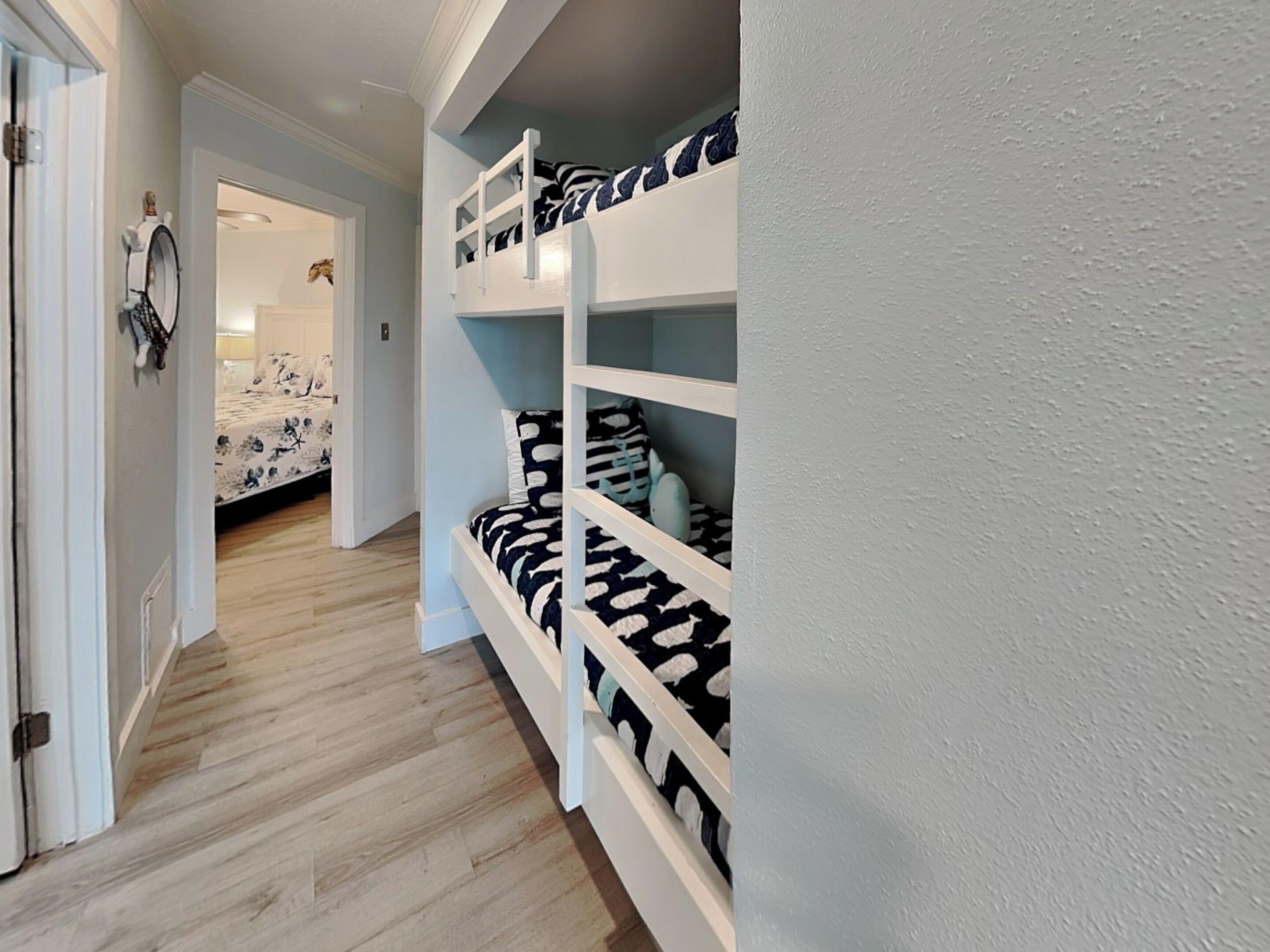 Un pasillo con una cama litera, suelo de madera y una puerta que da a una habitación con cama.