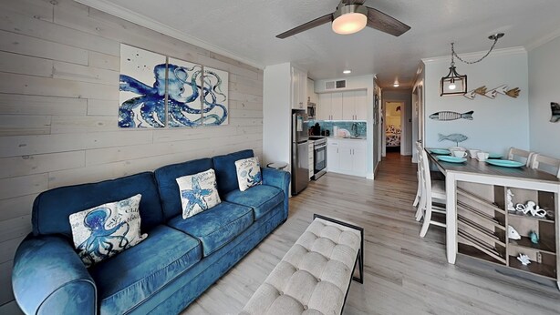 A modern living room with a blue sofa, a bench, a dining table with chairs, a kitchen in the background, and a ceiling fan. - Victorian 9124 - Good Times & Tan Lines (Galveston)