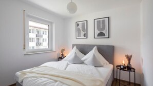 Quarto com cama, janela, duas obras de arte na parede e mesa de cabeceira com abajur.