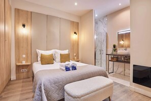 Ein modernes Schlafzimmer mit Bett, Nachttischen, einer Bank, einem Badezimmer mit Waschbecken und Spiegel sowie einem Kamin.