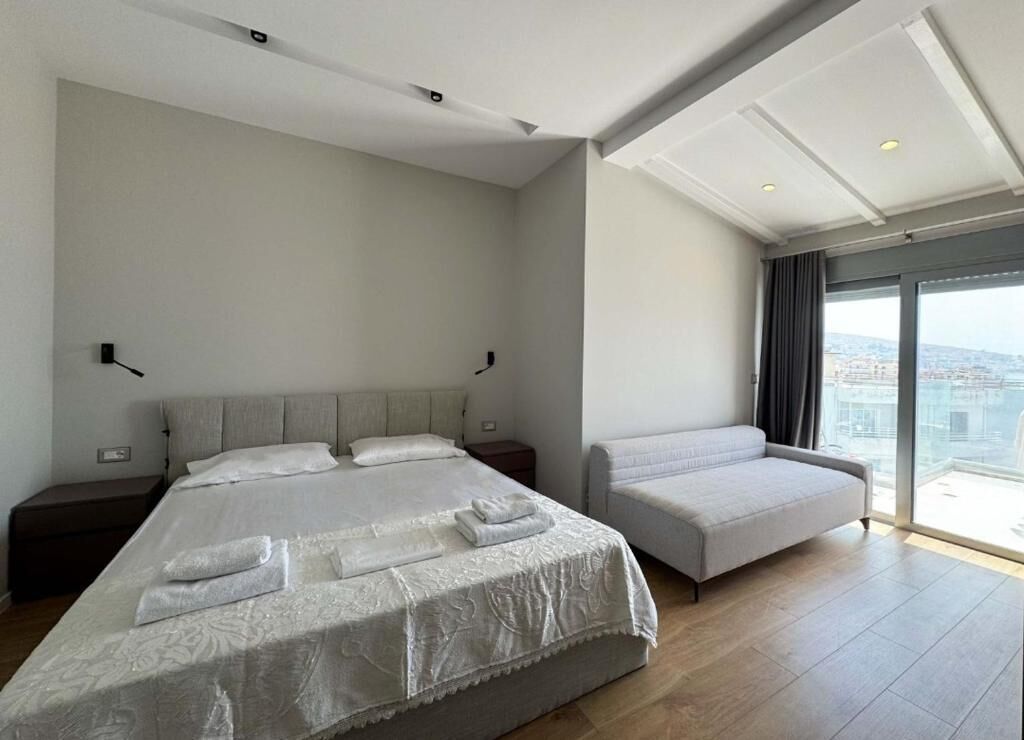 A modern hotel room with a large bed, a sofa, a view of the city, and a balcony.
