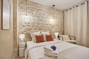 A bedroom with a stone wall, a bed with white bedding, two bedside lamps, and a chair. - Apartment 2/4 persons Eiffel Tower Invalides (Paris)