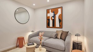 Een moderne woonkamer met een grijze bank, een ronde spiegel, een lijst met een abstract schilderij, een vaas met bloemen en een lamp op een bijzettafeltje.