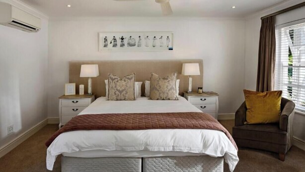 Un dormitorio de hotel con una cama grande, dos mesitas de noche con lámparas, una silla, un ventanal con cortinas y una obra de arte colgada en la pared.