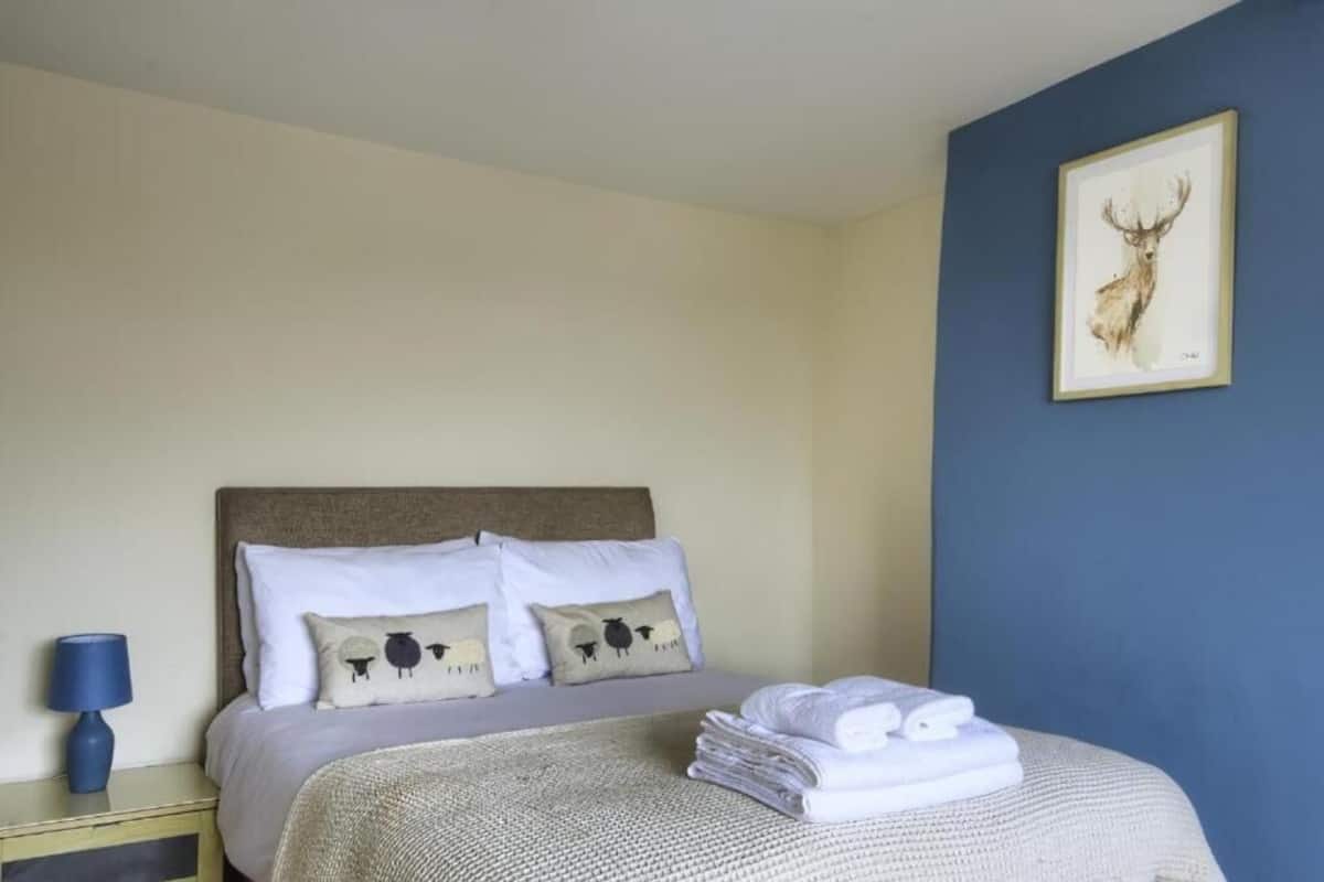 Un lit bien fait, avec du linge de lit blanc, une lampe bleue sur une table de chevet et un tableau représentant un cerf, encadré, accroché au mur.