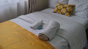 Een net opgemaakt bed met witte lakens, een gele deken en twee opgerolde handdoeken.