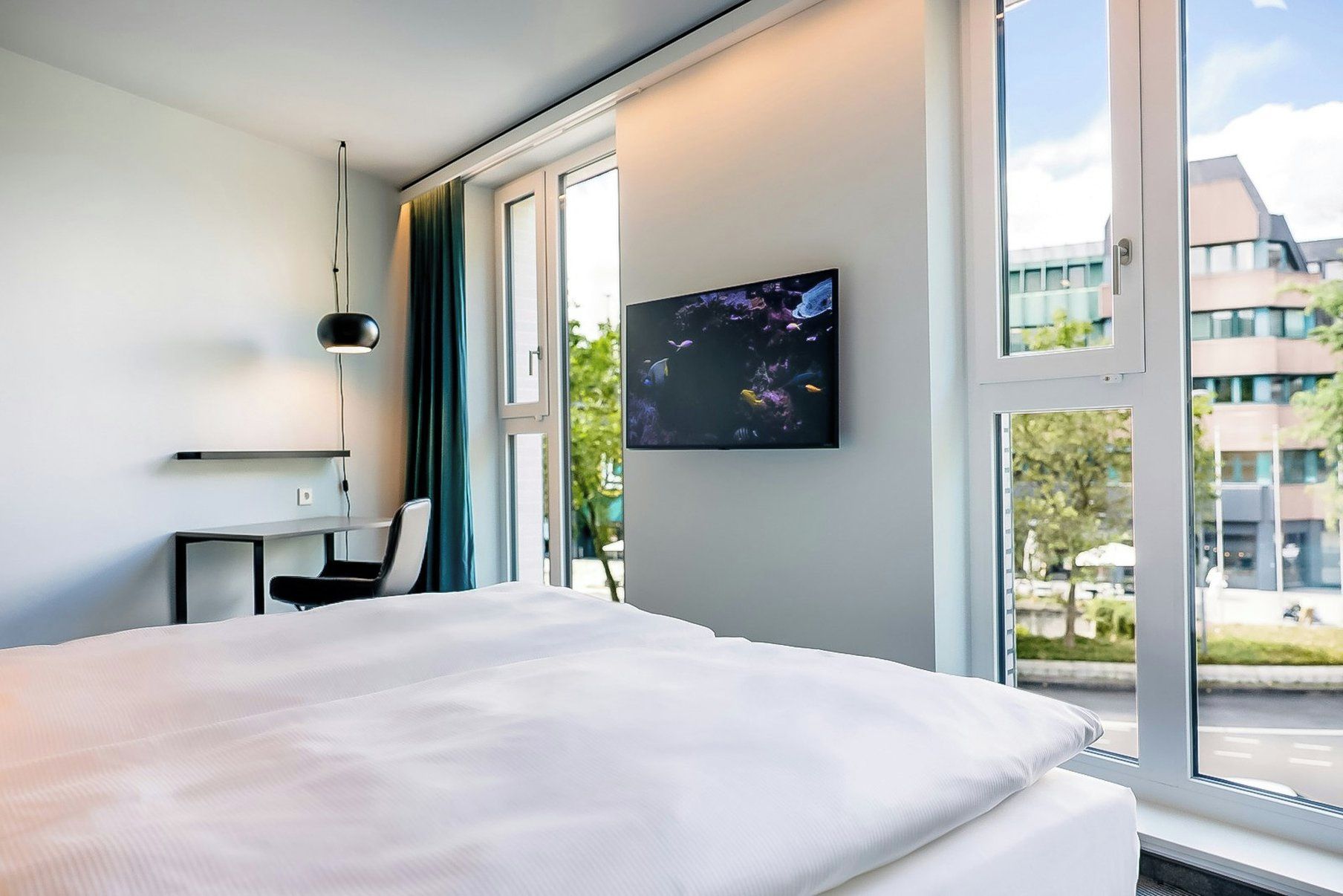A hotel room with a bed, a desk, a chair, a TV on the wall, and a window with a view of buildings and trees.