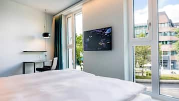 A hotel room with a bed, a desk, a chair, a TV on the wall, and a window with a view of buildings and trees.