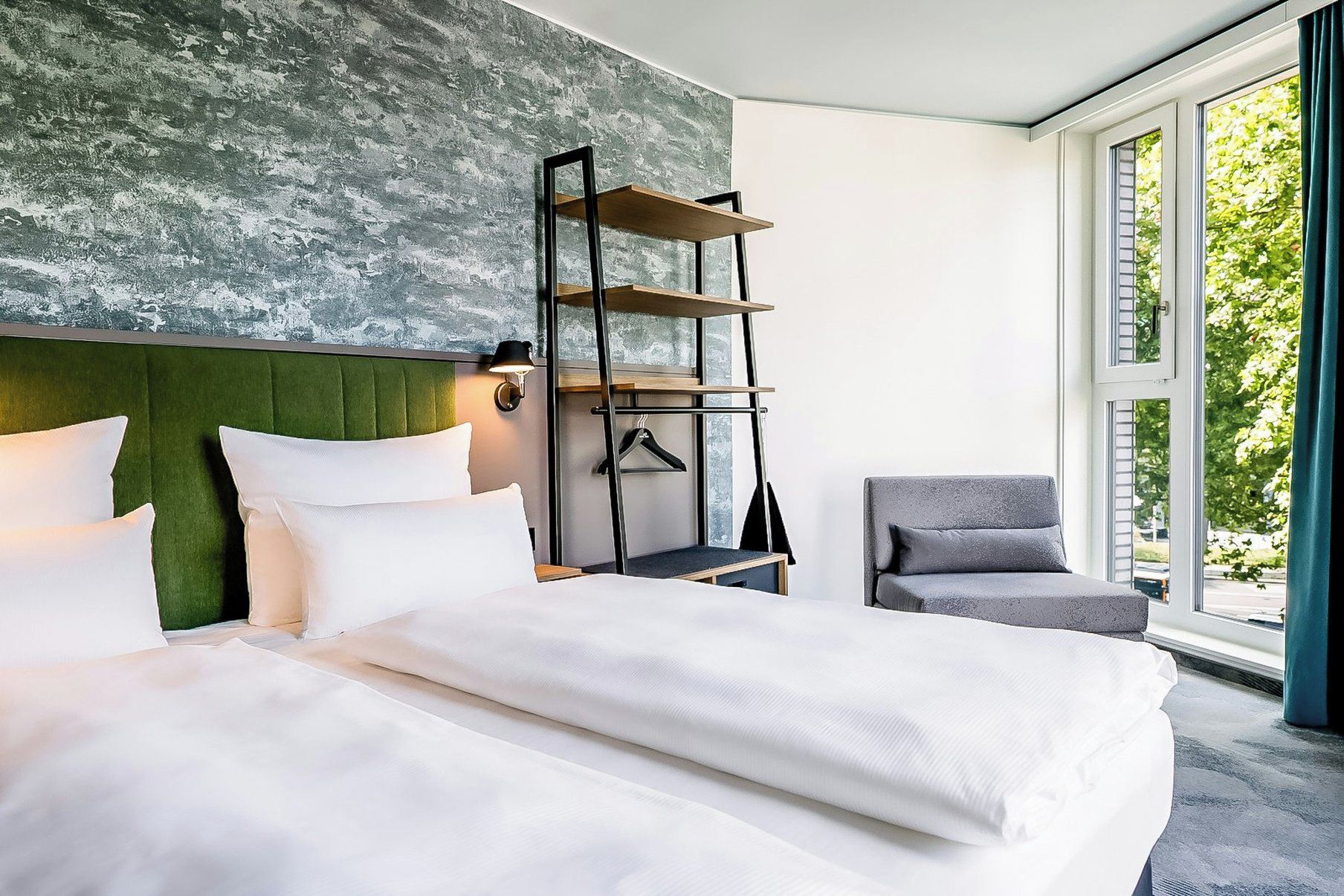 Une chambre d’hôtel avec un lit, une chaise, une étagère fixée au mur et une fenêtre donnant sur un paysage verdoyant.