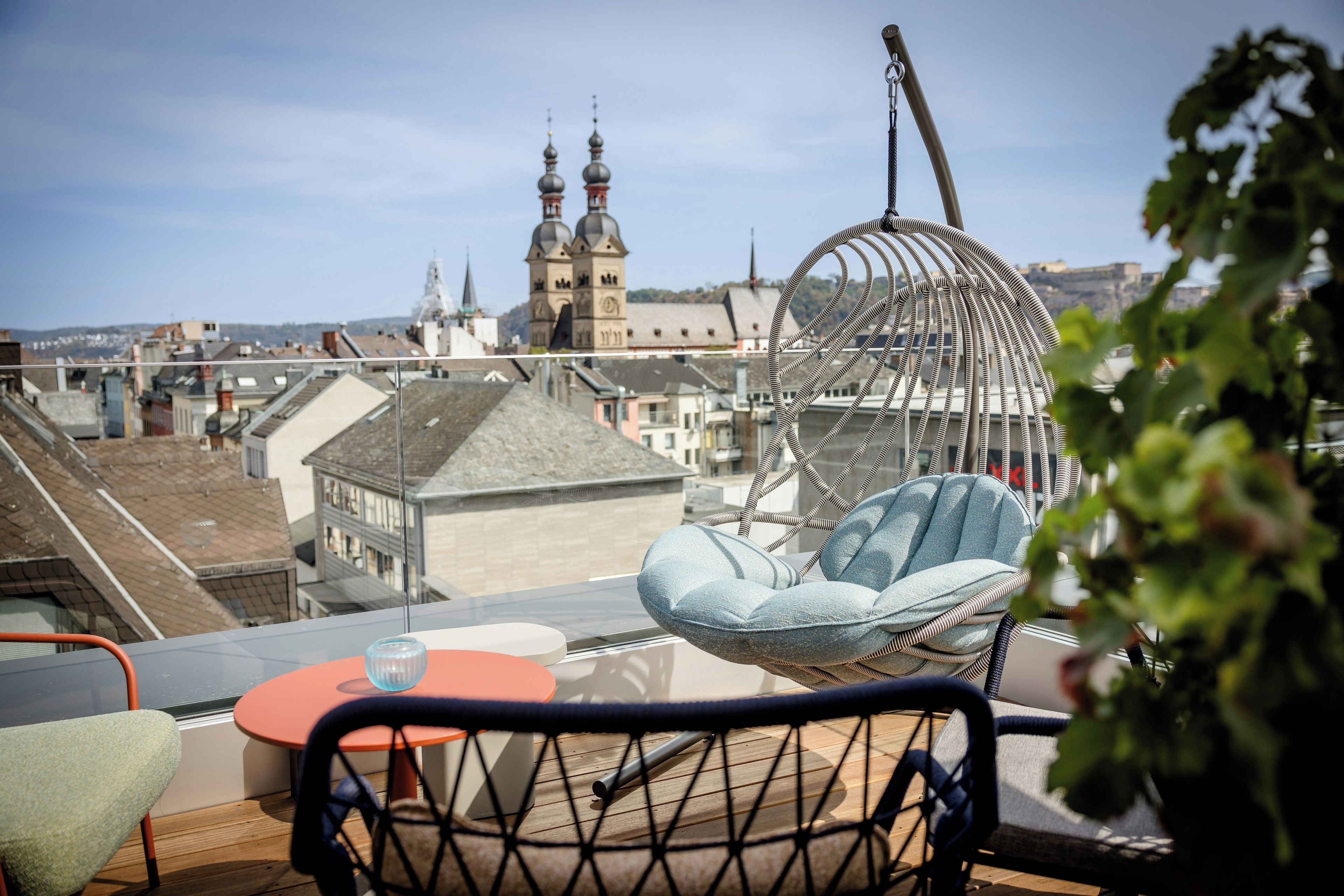 Ein Balkon mit einer Hängematten-Stuhlsitzgelegenheit, einem Tisch mit einem Glas und Blick auf eine Skyline mit einem Turm.