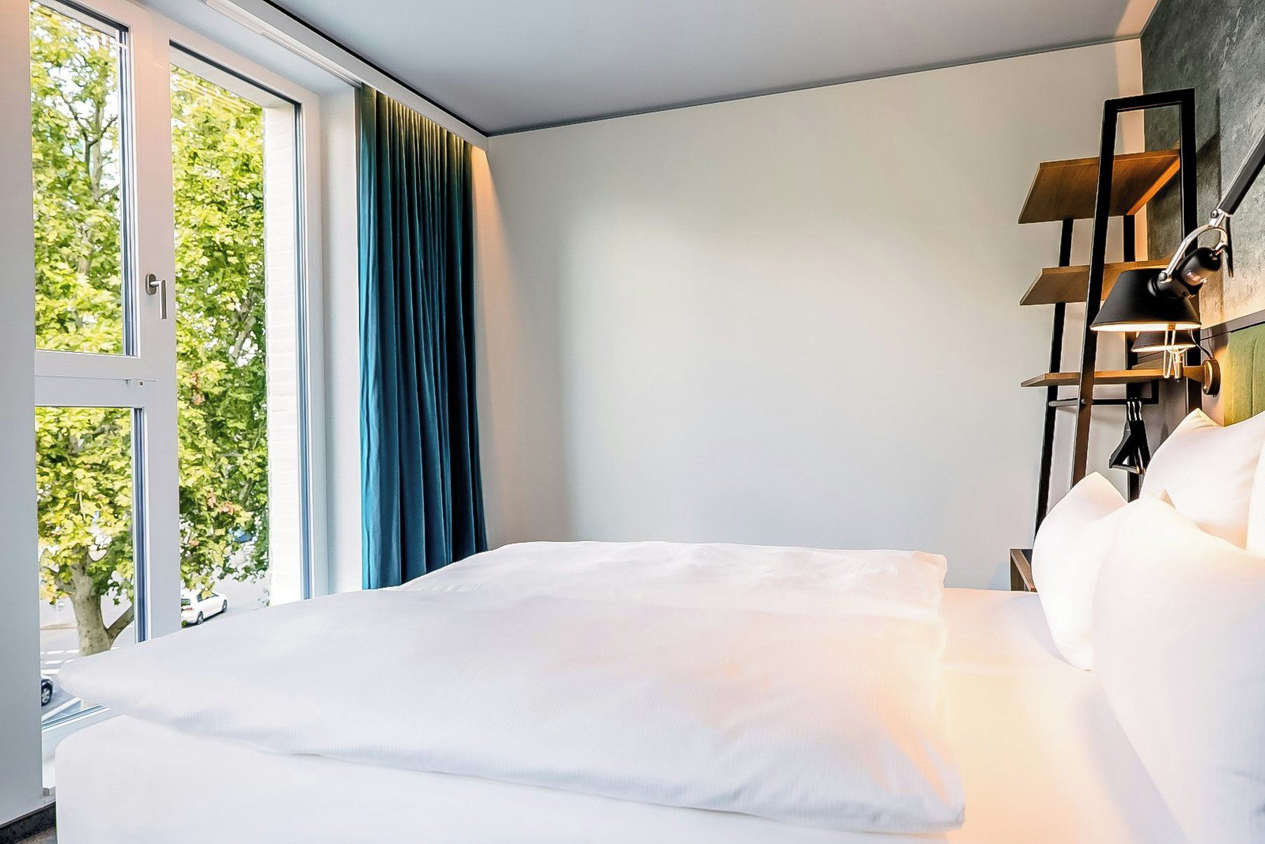 Una habitación de hotel con cama, una ventana con cortinas y una lámpara fijada a la pared.