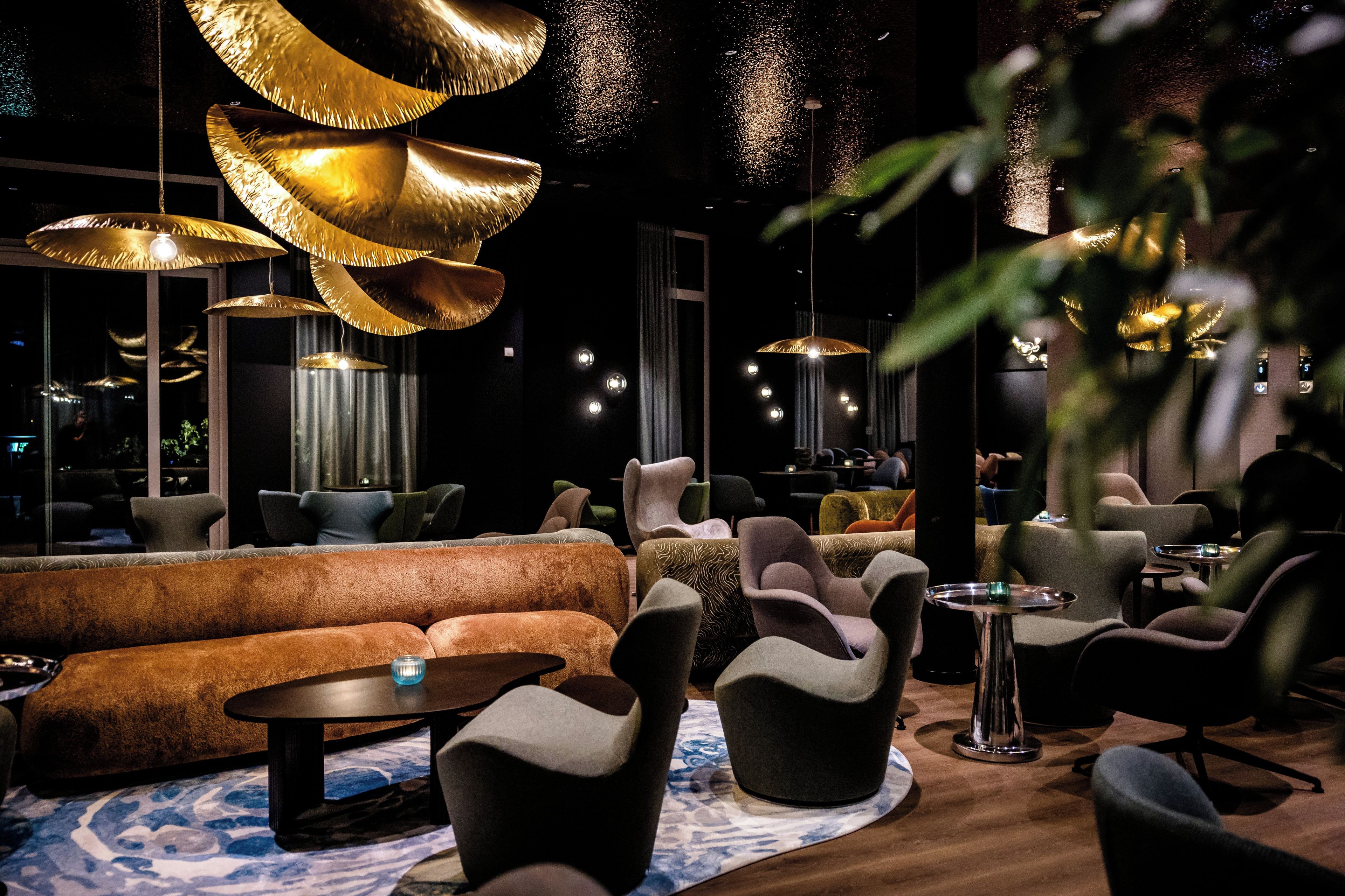 A modern lounge area with sofas, chairs, and tables, featuring unique pendant lighting and a patterned rug.