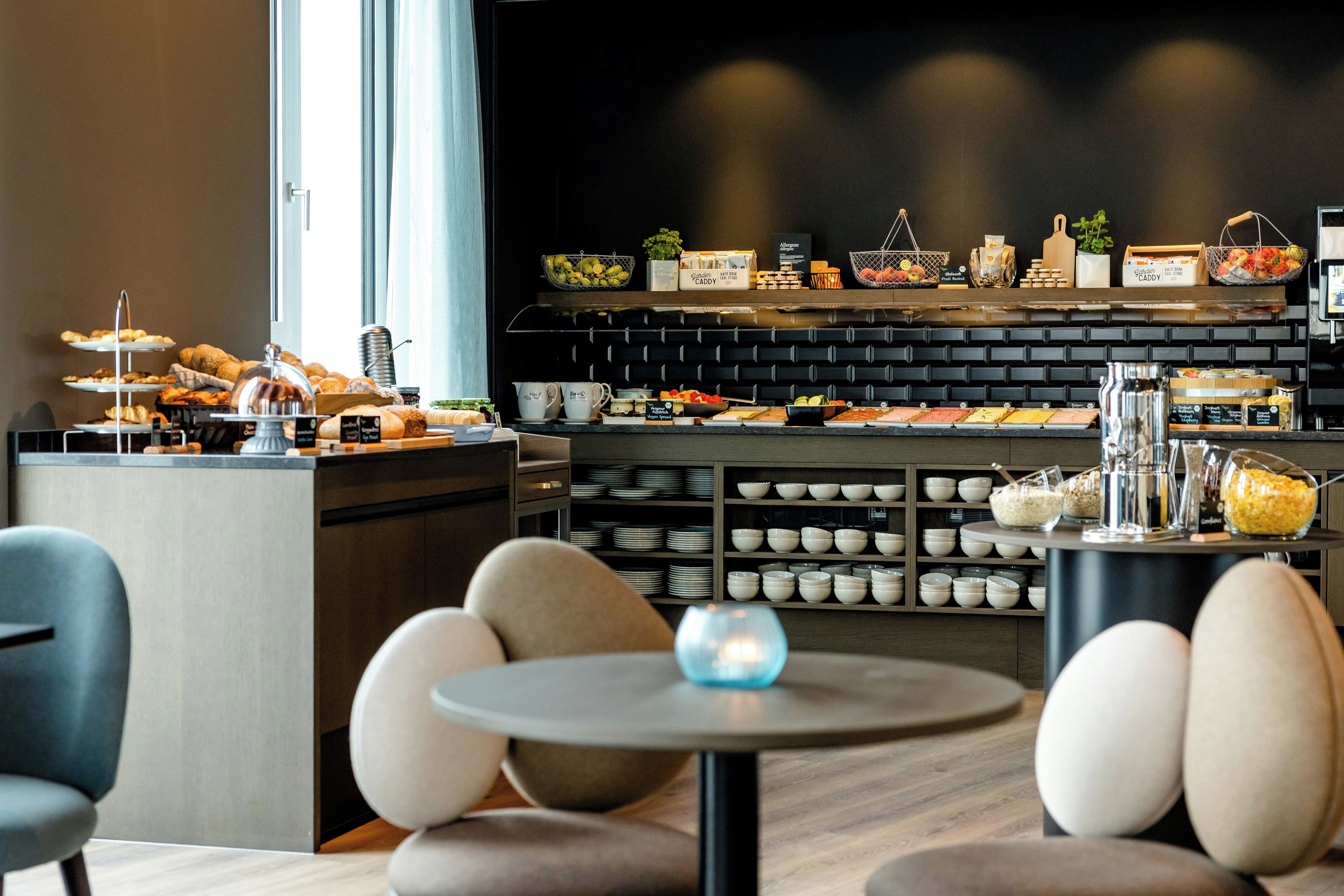 Interior moderno de una cafetería con una barra mostrando pasteles y bebidas, un área de mesas redondas y una pared con estantes llenos de tazones y platos.