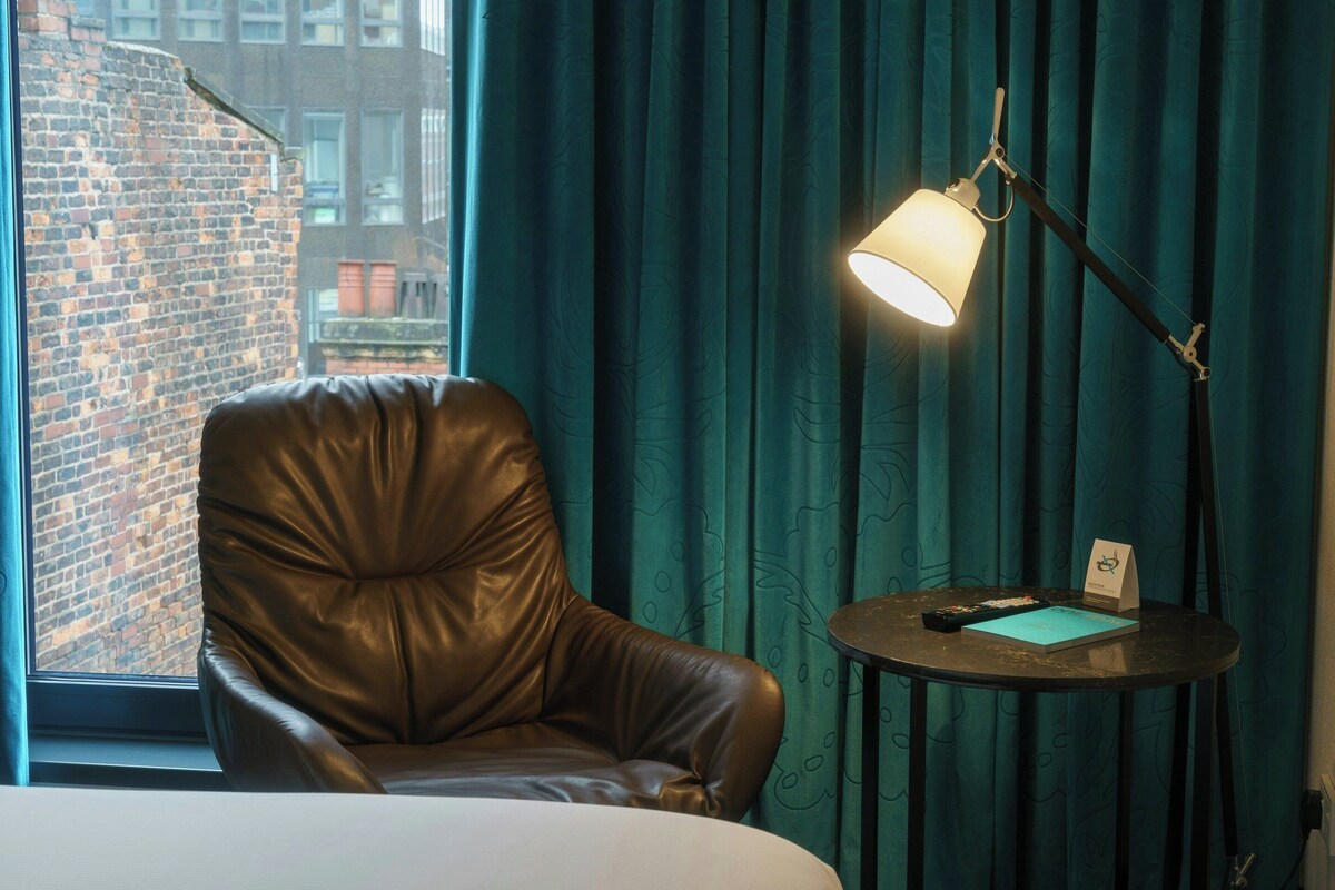 A leather chair, a bedside table with a lamp, and a view of a brick building through a window.