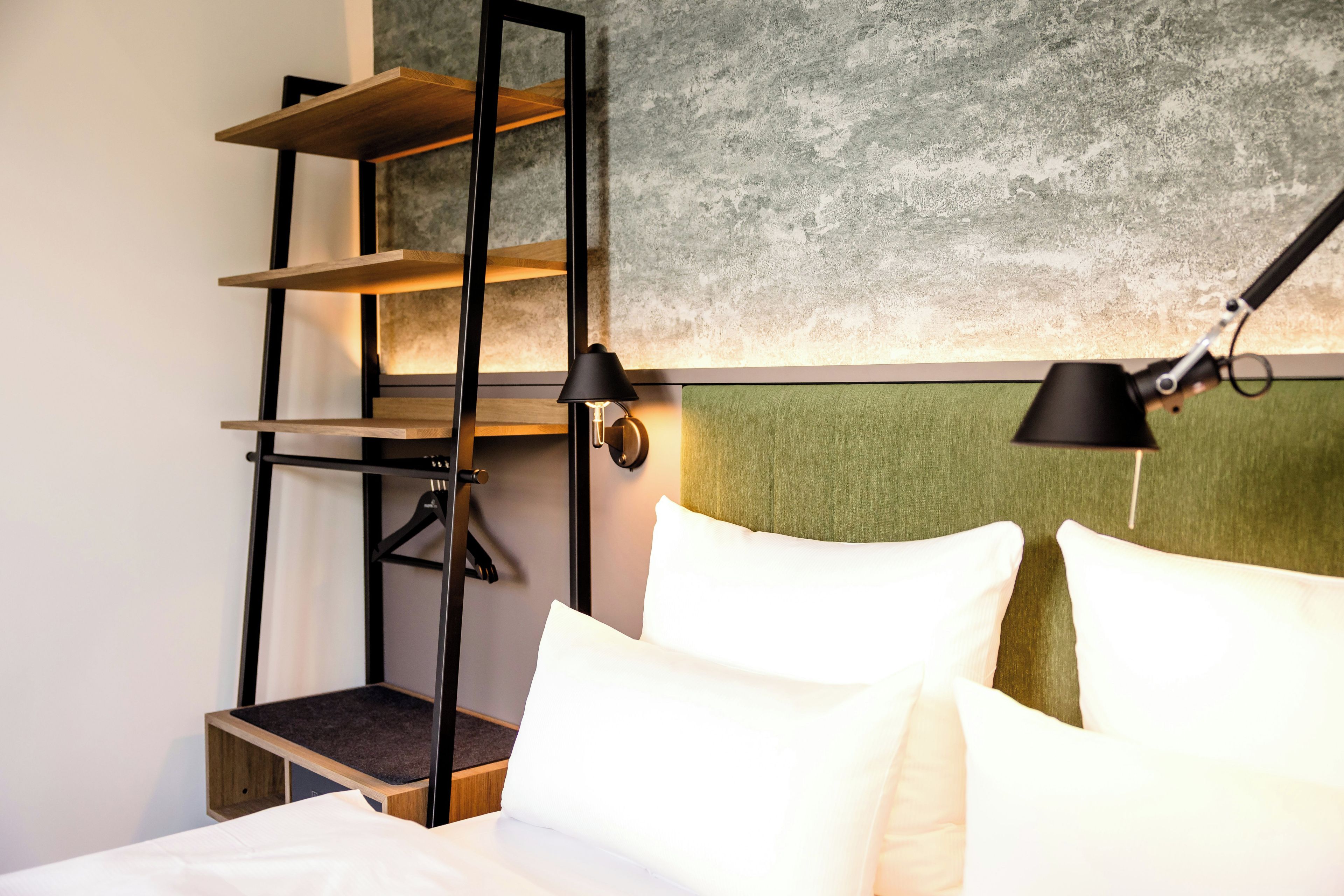 Una habitación de hotel con una cama, cabecera, estantería y dos lámparas fijadas a la pared.